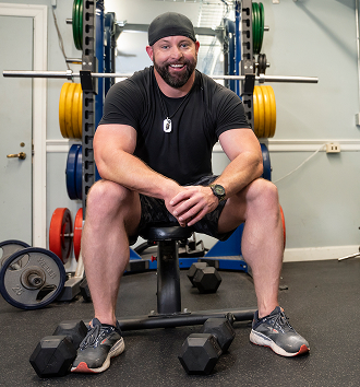 male Veteran in black shirt and shorts sitting on a bench press at the gym