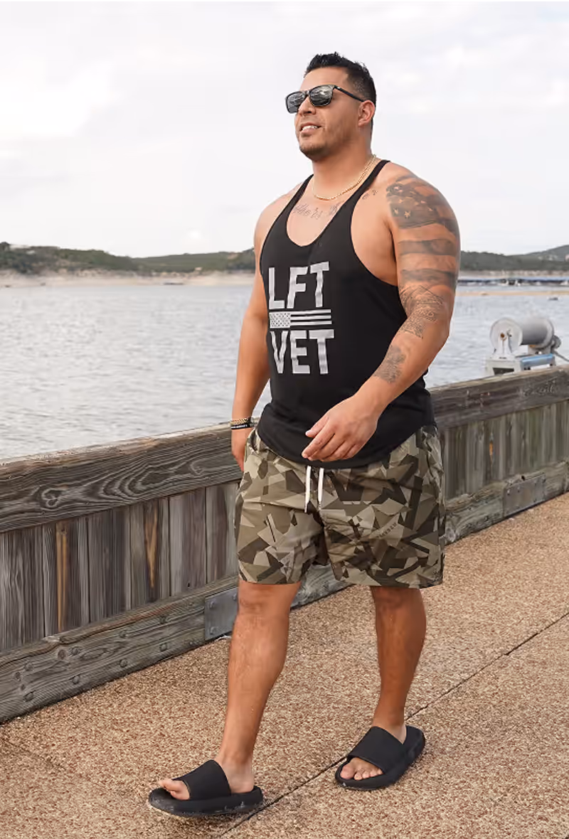 Man in black shirt, camo shorts and sunglasses walking on a pier with water in the background